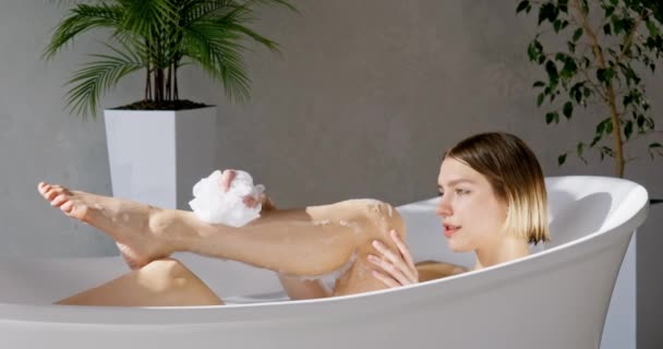 Sexy junge Frau wäscht Körper sanft und ruht sich in weißer bequemer Badewanne aus. Seitenansicht einer entspannten Frau mit Haarschnitt beim Baden, vor grauem Wandhintergrund, in Bewegung. Selbst- und Körperpflegekonzept. - Filmmaterial, Video