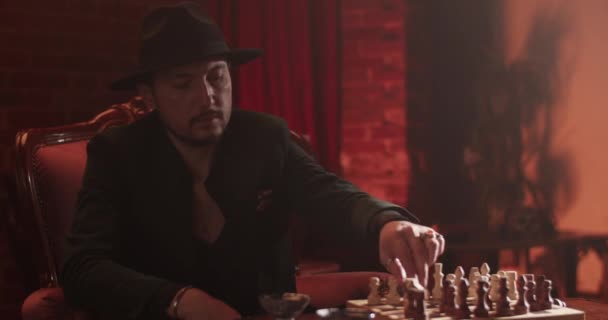Zelfverzekerde volwassen man met hoed en pak die schaakbordspel speelt met een anonieme vriend in de donkere kamer thuis - Video