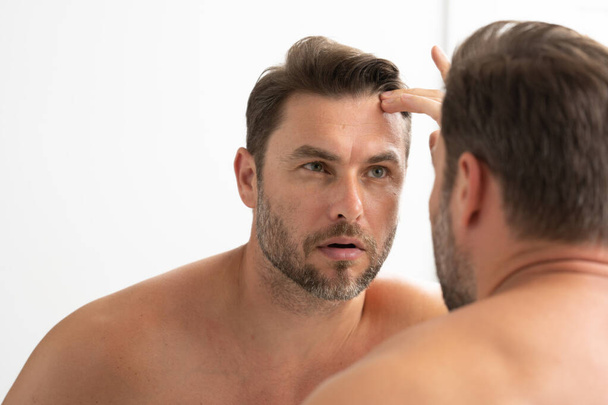 sexy man met stoppels kijkt naar de rimpels op zijn gezicht. Close-up man kijkt in spiegel, gevoelige huid, cosmetologie behandeling. Huidverzorging. vrolijk, viriel, mannelijk, aantrekkelijk, naakt, ongeschoren, knap - Foto, afbeelding