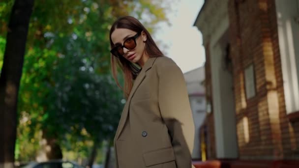 Gyönyörű lány divatos dzsekiben és napszemüvegben pózol a kamera előtt. Fiatal nő elegáns ruhában és szemüvegben sétál a város utcáján.. - Felvétel, videó