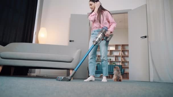 Uma jovem europeia em roupas acolhedoras em casa limpa o tapete do cabelo do cão de um terrier Yorkshire dourado com um aspirador de pó sem fio no apartamento - Filmagem, Vídeo