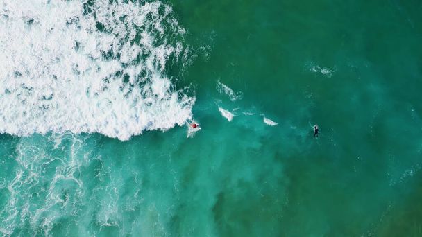 Top view surfingowiec pływanie w turkusowym basenie ocean piękny weekend letni. Piankowe fale burzowe toczące się do tropikalnej plaży. Strzał z powietrza nieznane osoby surfowanie w wodzie morskiej niebieski zwolniony ruch. - Zdjęcie, obraz