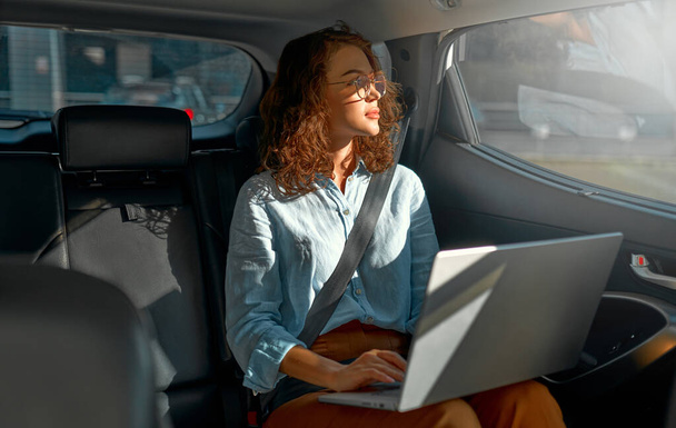 Young business woman with a modern laptop sitting in the back seat of luxury car. Successful startup, distance working, female boss concept. - Photo, Image