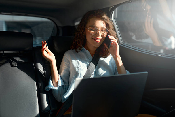 Attraente donna d'affari che lavora sul suo computer portatile sul sedile posteriore di una macchina e parla al telefono sulla strada per l'incontro
. - Foto, immagini