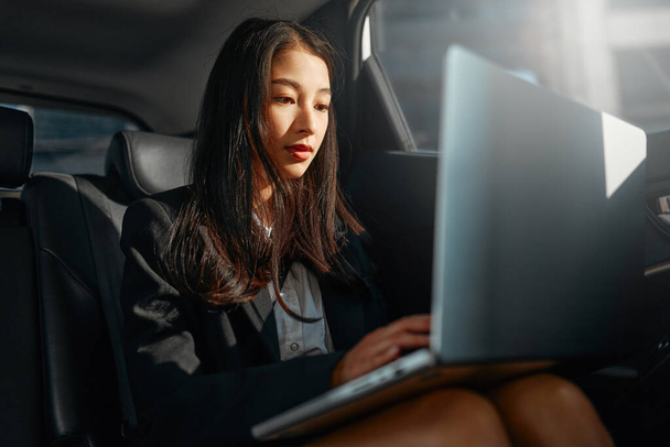 Fiatal ázsiai üzletasszony egy modern laptoppal a luxusautó hátsó ülésén. Sikeres startup, távmunka, női főnök koncepció. - Fotó, kép