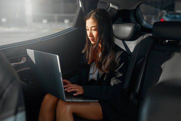 Young asian business woman with a modern laptop sitting in the back seat of luxury car. Successful startup, distance working, female boss concept. - Photo, Image