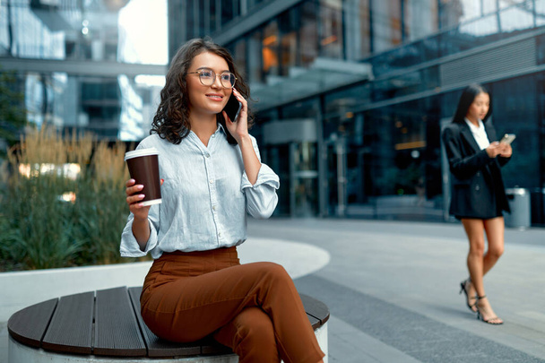 Eine junge schöne Geschäftsfrau trinkt Kaffee und benutzt ein Smartphone, während sie auf der Straße vor dem Hintergrund von Geschäftszentren sitzt. - Foto, Bild