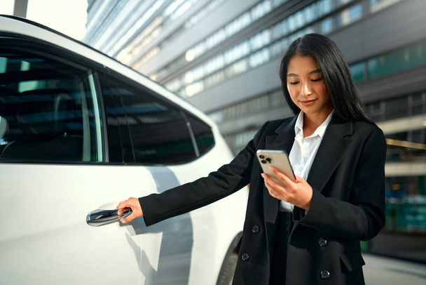 Piękny młody szczęśliwy azjatycki biznes kobieta w czarnym garniturze stoi w pobliżu jej nowego samochodu i za pomocą smartfona. Kupuję i wynajmuję samochód. Podróże, turystyka, rekreacja. - Zdjęcie, obraz
