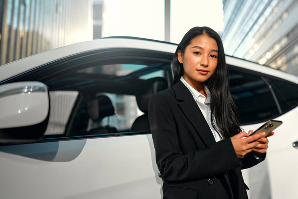 Krásná mladá šťastná asijská obchodní žena v černém obleku stojí v blízkosti svého nového auta a pomocí smartphonu. Kupuje a pronajímá auto. Cestování, cestovní ruch, rekreace. - Fotografie, Obrázek