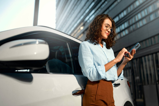 Красива молода щаслива кавказька бізнес-леді, що стоїть біля своєї нової машини і використовує смартфон. Купівля та оренда автомобіля. Подорожі, туризм, відпочинок. - Фото, зображення