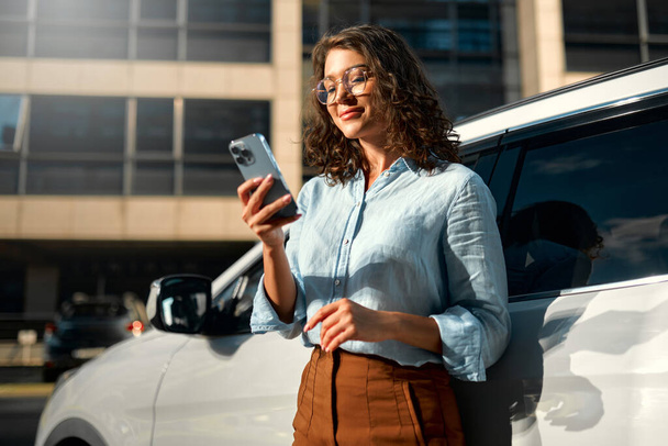 Piękna młoda, szczęśliwa, biała kobieta biznesu, stojąca blisko swojego nowego samochodu i używająca smartfona. Kupuję i wynajmuję samochód. Podróże, turystyka, rekreacja. - Zdjęcie, obraz