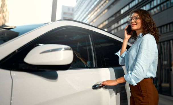 Kaunis nuori onnellinen valkoihoinen liikenainen seisoo lähellä uutta autoaan ja käyttää älypuhelinta. Auton ostaminen ja vuokraaminen. Matkailu, matkailu, virkistys. - Valokuva, kuva