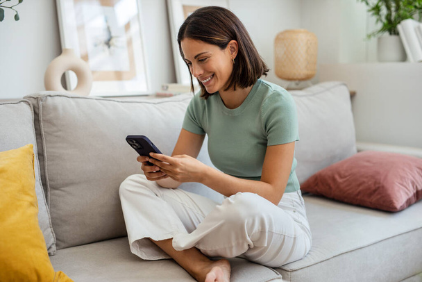 Shot of smiling young woman using her mobile phone while sitting on sofa at home - Foto, imagen