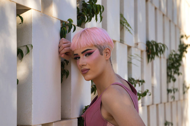 Menino gay jovem com cabelo rosa e maquiagem encostada a uma parede branca com buracos retangulares em estilo moderno. O rapaz faz expressões diferentes. Conceito de igualdade e direitos LGBTQ - Foto, Imagem
