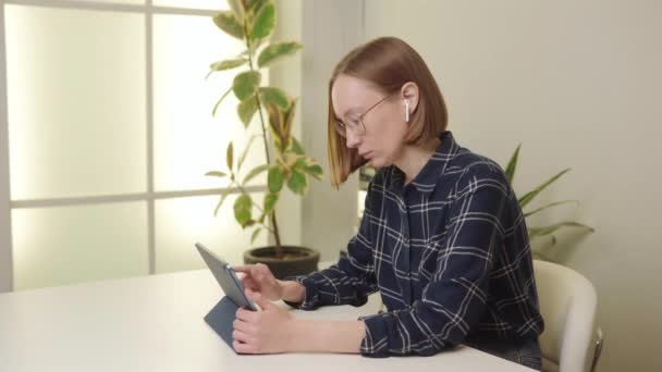 Kareli gömlekli bir kadın, arka planda saksı bitkisi olan beyaz bir ofis masasında tablet kullanıyor. Yüksek kalite 4k görüntü. - Video, Çekim