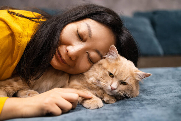 Güzel Asyalı kadın, turuncu kedinin yanına uzanıp kedisinin çenesini kaşıyarak onu rahatlatır ve yatakta mutlu eder.. - Fotoğraf, Görsel