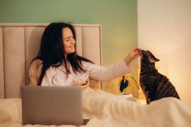 Frau im Schlafanzug liegt im Bett und füttert eine Katze - Foto, Bild