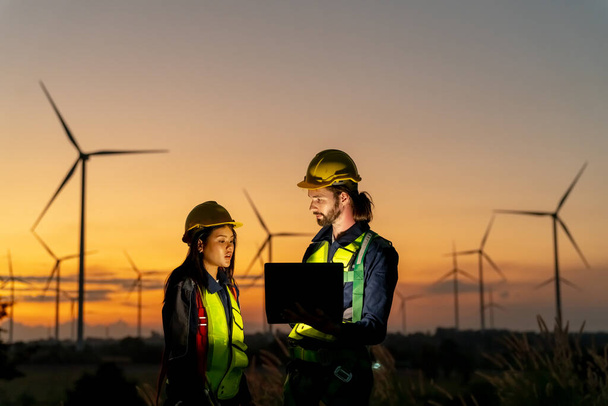 Δύο πολυφυλετικές βιομηχανικές εργάτριες που εξετάζουν ανεμογεννήτριες. Αγροτική περιοχή με οικολογική εκμετάλλευση παραγωγής καθαρής πράσινης ενέργειας - Φωτογραφία, εικόνα