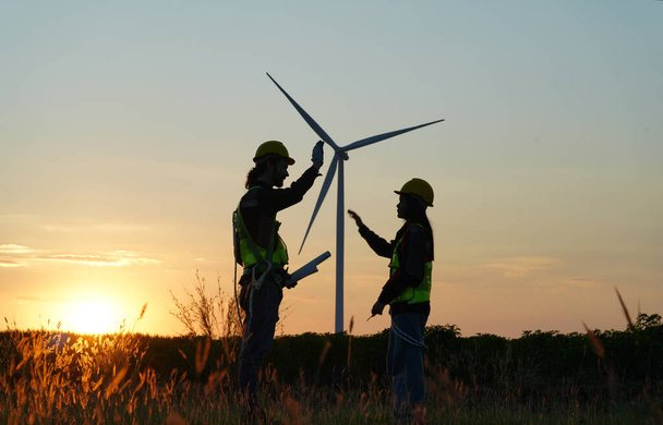 Kaksi monirotuista teollisuustyöntekijää tutkii tuulivoimaloita. Maaseutualue, jolla on ekologinen tuotantotila - puhdas vihreä energia - Valokuva, kuva