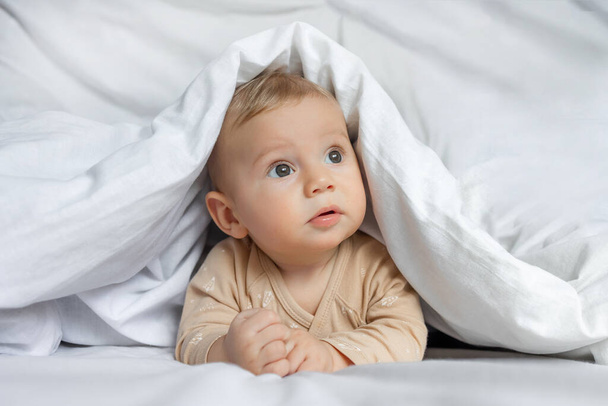 Χαριτωμένο μωρό παίζει κρύβεται κάτω από λευκή κουβέρτα στο σπίτι. - Φωτογραφία, εικόνα