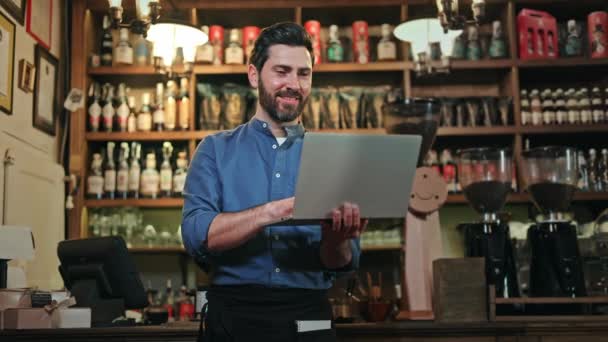 モダンなラップトップを手に持っているブルーシャツのハッピーマネージャーは,笑顔ながらカフェについて肯定的なレビューをチェックします. 小規模ビジネスを促進するための技術デバイスとネットを使用して有能な男性従業員. - 映像、動画