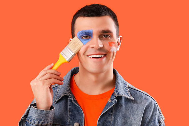 Молодой человек с раскрашенным лицом и кистью на оранжевом фоне, крупным планом - Фото, изображение
