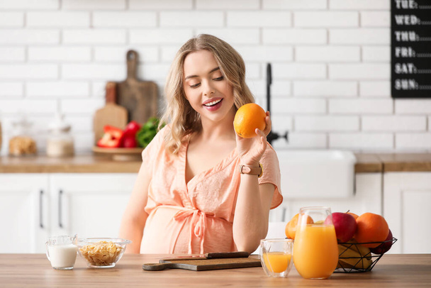 Mladá těhotná žena s pomerančem u stolu v kuchyni - Fotografie, Obrázek