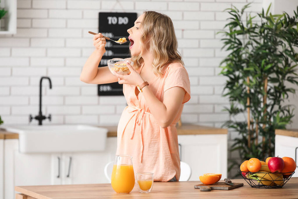 Молодая беременная женщина ест кукурузные хлопья во время завтрака на кухне - Фото, изображение