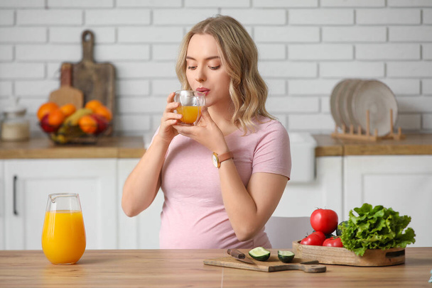 Νεαρή έγκυος γυναίκα πόσιμο χυμό στο τραπέζι στην κουζίνα - Φωτογραφία, εικόνα