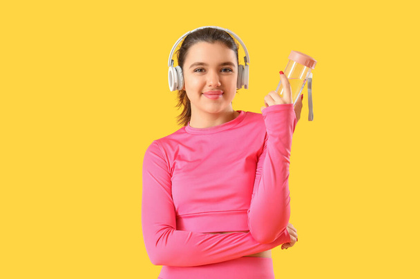 Young woman in headphones with sports water bottle on yellow background - Zdjęcie, obraz