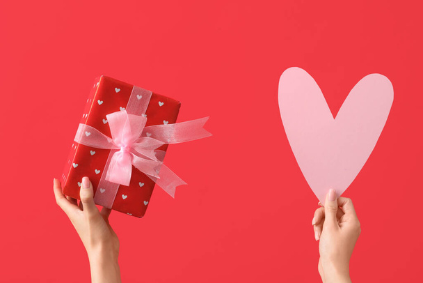 Female hands with paper heart and gift box on red background. Valentine's Day celebration - Photo, Image