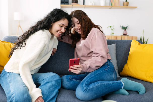 Two friendly high school student female friends relaxing together on sofa having fun using mobile phone. Friendship and technology lifestyle concept - Photo, Image