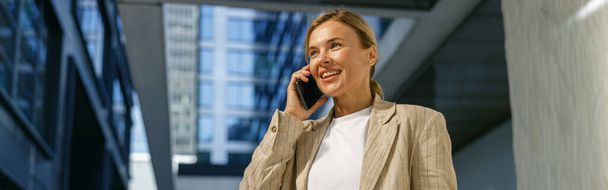 Positive female freelancer holding laptop and talking phone standing on modern building background - Photo, Image