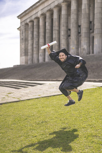 Man recently graduated, dressed in cap and gown, with her degree in her hand, jumping, on the university campus. Very happy expression, achievement, beginning of a new stage - Photo, Image