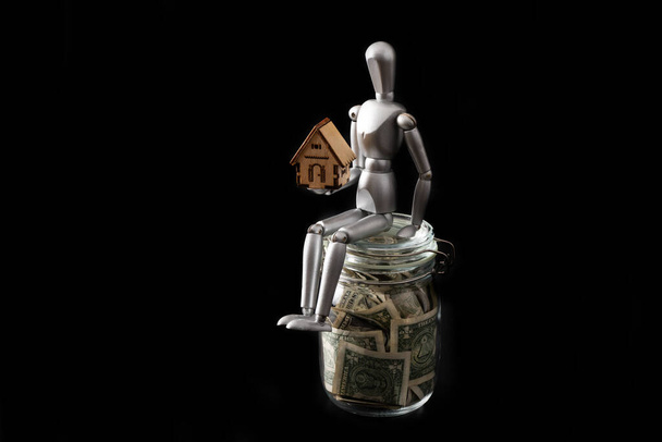 Üvegedény 1 USA dollár bankjegyekkel, fából készült férfi és szimbolikus ház fekete alapon - Fotó, kép