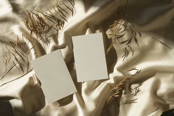 Αισθητικό bohemian branding πολυτελείας ή προσκλητήριο πρότυπο καρτών. Κενό φύλλο κάρτας χαρτιού με κενό mock up χώρο αντιγραφής, αποξηραμένα στελέχη γρασίδι σε τσαλακωμένο γυαλιστερό χρυσό φόντο μετάξι με σκιές φως του ήλιου - Φωτογραφία, εικόνα