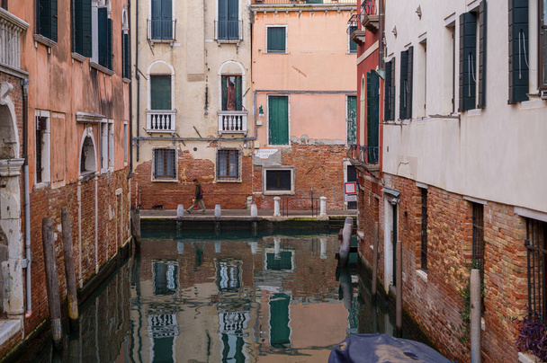 In an area of Venice (Italy) that is rarely visited by tourists, a man in a scarf and hat walks quickly with his small dog and is reflected in the nearby waterway. - Photo, Image