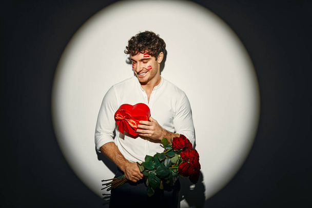 Hyvää ystävänpäivää. Nuori seksikäs mies, jolla on ruusukimppu ja lahja, jossa on jälkiä punaisesta huulipunasta kasvoillaan suukoista, seisoo valkoisella taustalla valokeilan valossa.. - Valokuva, kuva