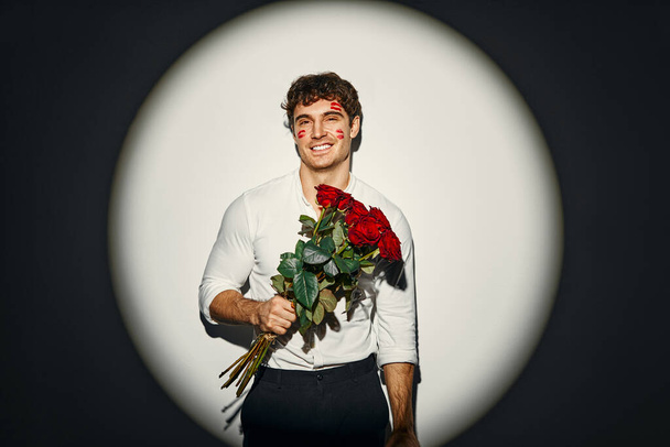 Feliz Dia dos Namorados. Homem sexy com um buquê de rosas com traços de batom vermelho na forma de lábios em seu rosto de beijos, de pé sobre um fundo branco sob a luz de um holofote. - Foto, Imagem