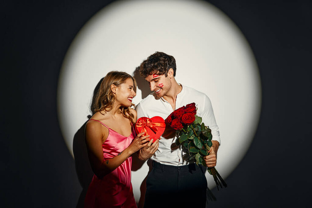 Χρόνια πολλά για του Αγίου Βαλεντίνου. Ζευγάρι ερωτευμένο στέκεται με ένα κουτί δώρου σε σχήμα καρδιάς και ένα μπουκέτο τριαντάφυλλα σε λευκό φόντο κάτω από το φως ενός προβολέα. - Φωτογραφία, εικόνα