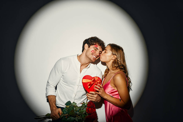 С Днем Святого Валентина. Влюбленная пара, стоящая с подарочной коробкой в форме сердца и букетом роз на белом фоне под светом прожектора. - Фото, изображение
