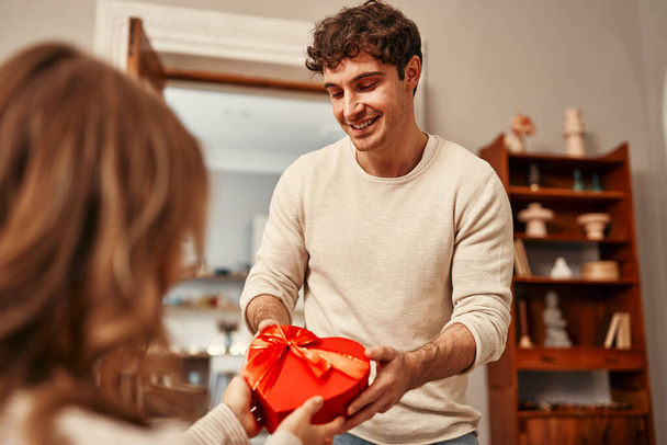 Feliz día de San Valentín. Un hombre le da una caja de regalo en forma de corazón a su amada mujer en la sala de estar en casa. Noche romántica juntos. - Foto, Imagen