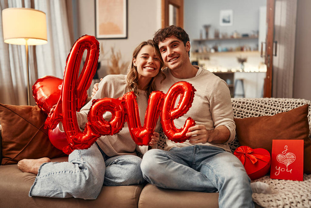 Feliz día de San Valentín. Pareja joven enamorada sosteniendo un globo con el amor de inscripción mientras está sentada en el sofá en la sala de estar en casa. Noche romántica juntos. - Foto, imagen