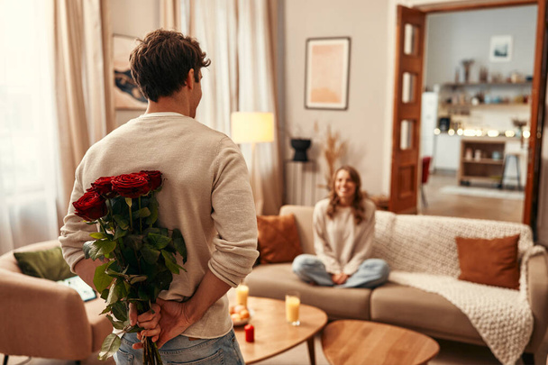 Šťastného Valentýna. Muž držící za zády kytici rudých růží pro svou milovanou ženu, která sedí doma na gauči v obývacím pokoji. - Fotografie, Obrázek