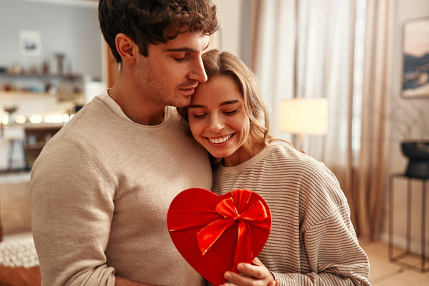 Boldog Valentin napot! Egy férfi odaad egy szív alakú ajándékdobozt szeretett nőjének a nappaliban, a nő gyengéden megöleli. Romantikus este együtt.. - Fotó, kép