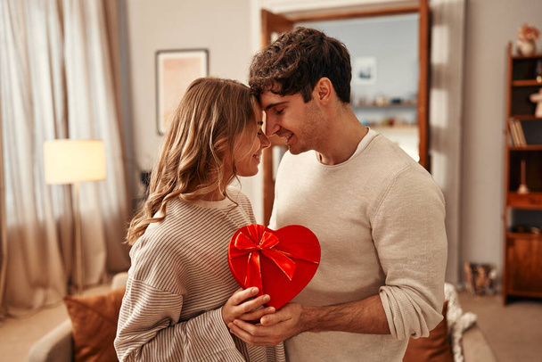 Alles Gute zum Valentinstag. Ein Mann schenkt seiner geliebten Frau im heimischen Wohnzimmer eine herzförmige Geschenkschachtel, die Frau umarmt ihn zärtlich. Romantischer gemeinsamer Abend. - Foto, Bild