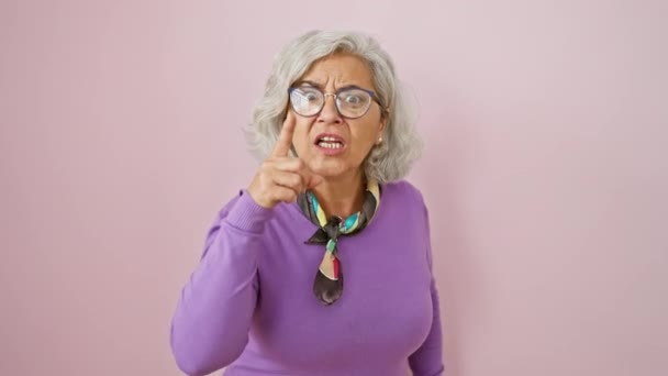Mujer furiosa de mediana edad, de pelo gris y con gafas, señalándote con el dedo acusándote frente a un fondo rosa aislado, rebosante de ira y frustración, claramente disgustada. - Metraje, vídeo