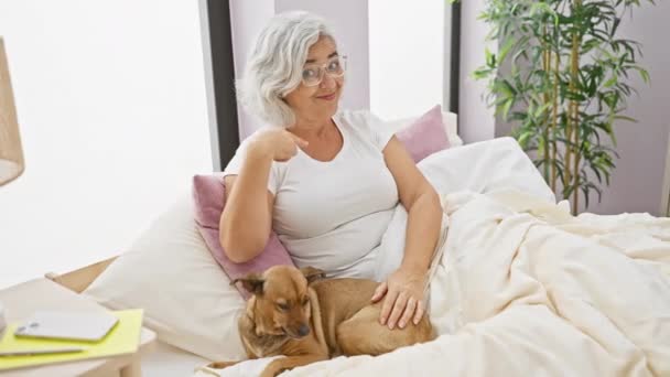 Vrolijke vrouw van middelbare leeftijd, met grijs haar, vrolijk wijzend naar zichzelf terwijl ze in pyjama op bed zat met haar trotse hond in de slaapkamer. - Video