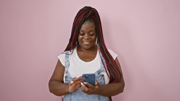 Selbstbewusste afrikanisch-amerikanische Frau, die freudig auf ihrem Smartphone SMS schreibt und über isolierten rosa Hintergrund lächelt - Filmmaterial, Video
