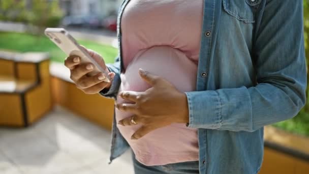 Mladá těhotná žena dotýká břicho pomocí smartphone v parku - Záběry, video